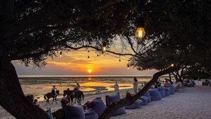 A beach scene with seating areas under trees, a sunset in the background, and people and horses on the sand. - Pearl Sunset Resort (Gili Trawangan)