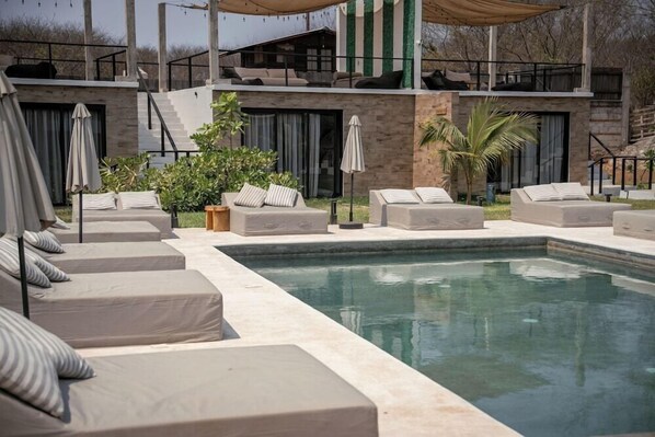 A modern outdoor pool area with multiple lounge chairs and umbrellas, surrounded by a stone building with a staircase and a view of the surrounding landscape. - Bora Boutique (Santa María Colotepec)