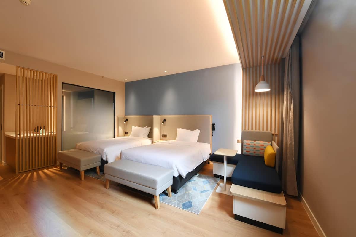 A modern hotel room with two beds, a desk, a bench, and a small table.