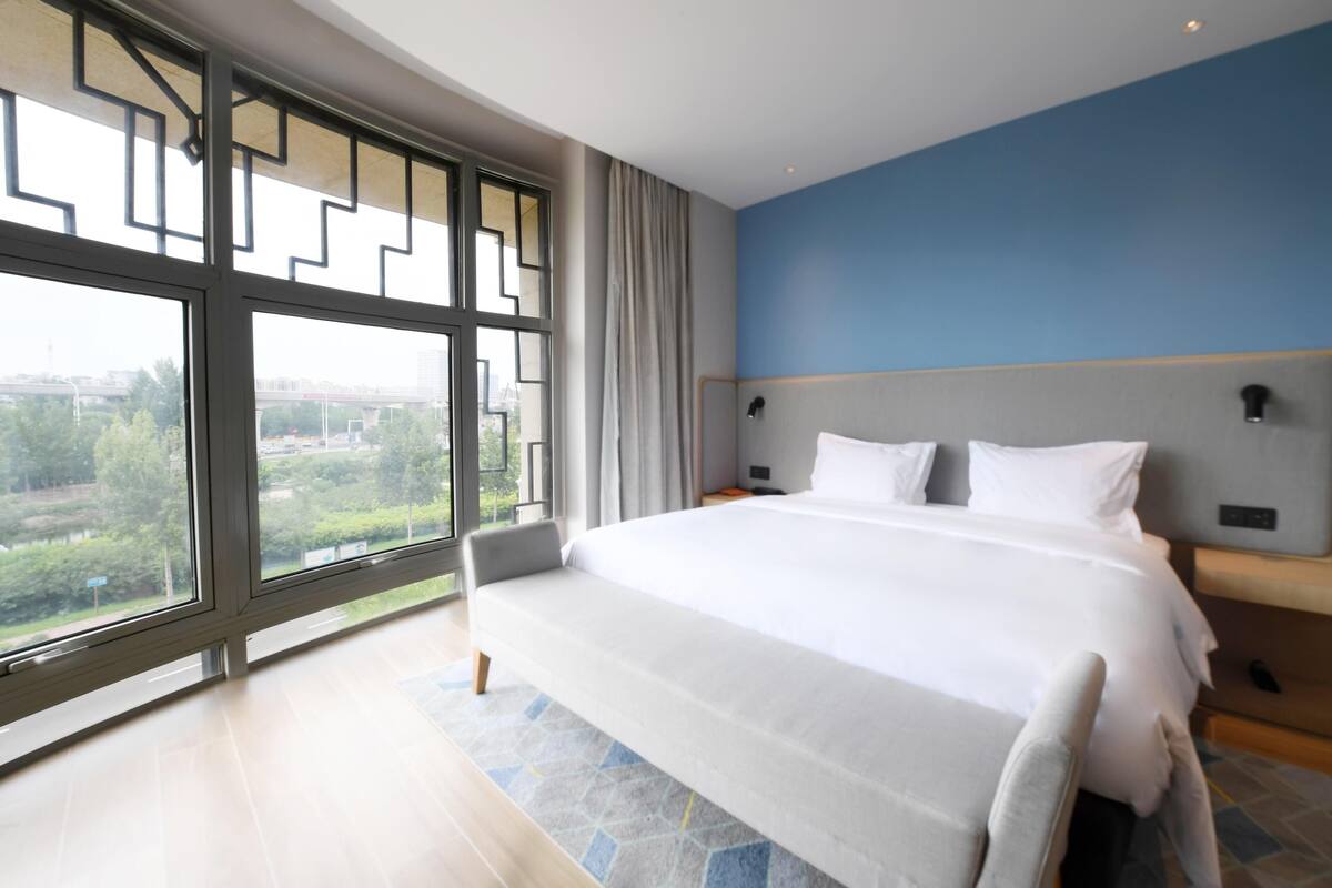 Habitación de hotel moderna con una cama grande, vistas a zonas verdes a través de ventanales de gran altura y una pared de acento azul.