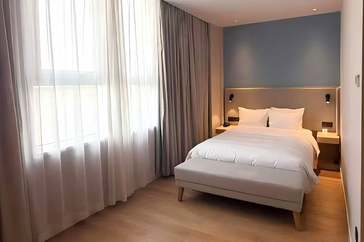 Habitación de hotel con una cama grande, una mesita de noche, una lámpara de pared y cortinas translúcidas.