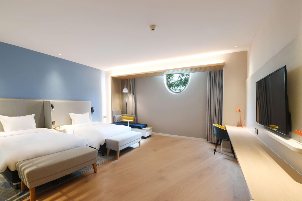 Ein Hotelzimmer mit zwei Betten, einem Schreibtisch, einem Fernseher und einem runden Fenster.