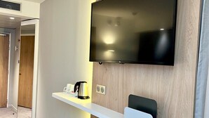 Une chambre d’hôtel moderne dotée d’un téléviseur à écran plat fixé au mur, d’une étagère blanche avec une bouilloire et une machine à café, et d’un mur d’accent en bois.