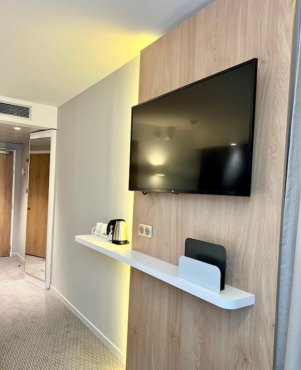 Une chambre d’hôtel moderne dotée d’un téléviseur à écran plat fixé au mur, d’une étagère blanche avec une bouilloire et une machine à café, et d’un mur d’accent en bois.