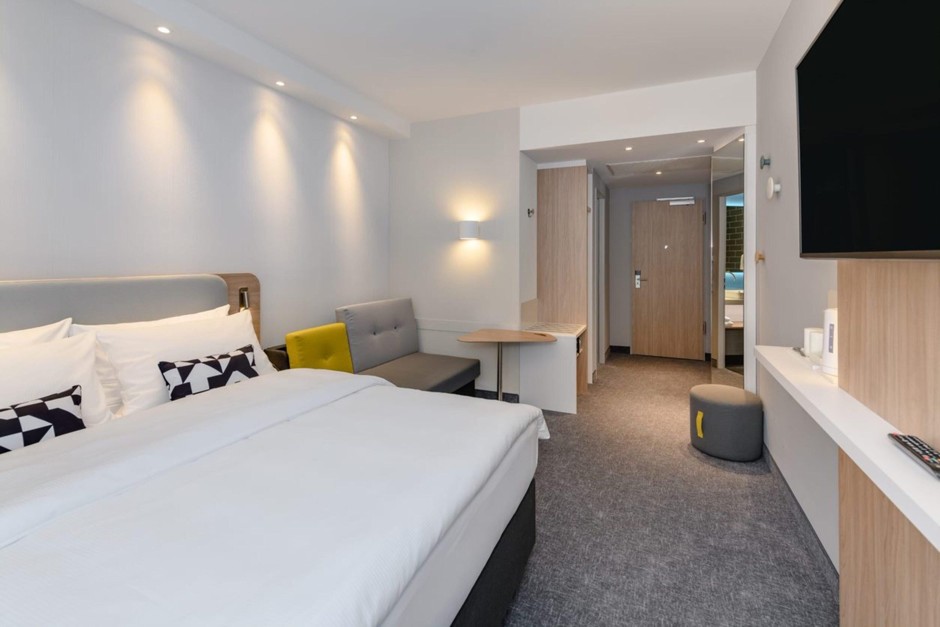 A modern hotel room with a large bed, a small desk, a seating area, and a TV mounted on the wall.