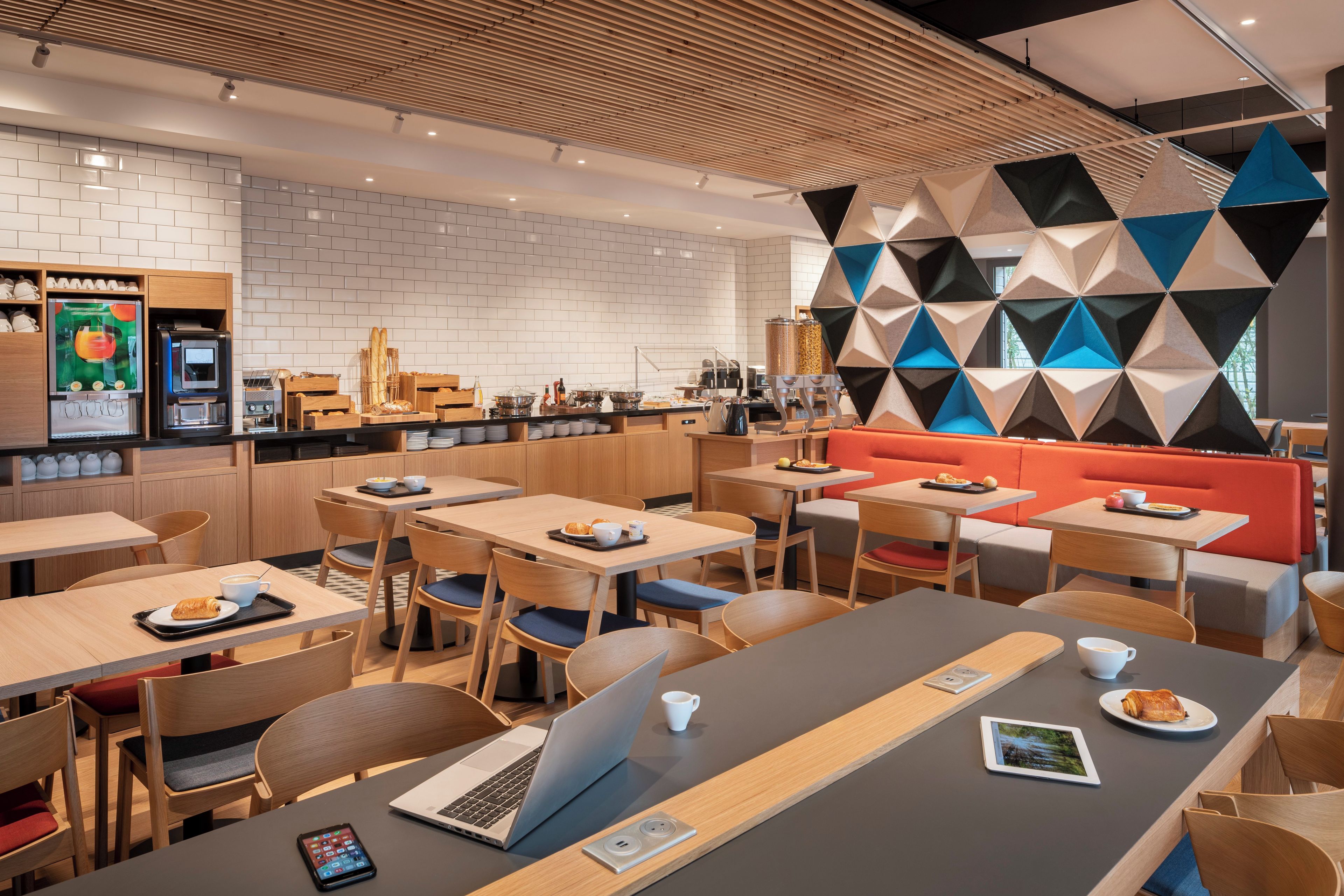 A modern cafe with wooden tables and chairs, a counter with a coffee machine, and a colorful geometric wall.