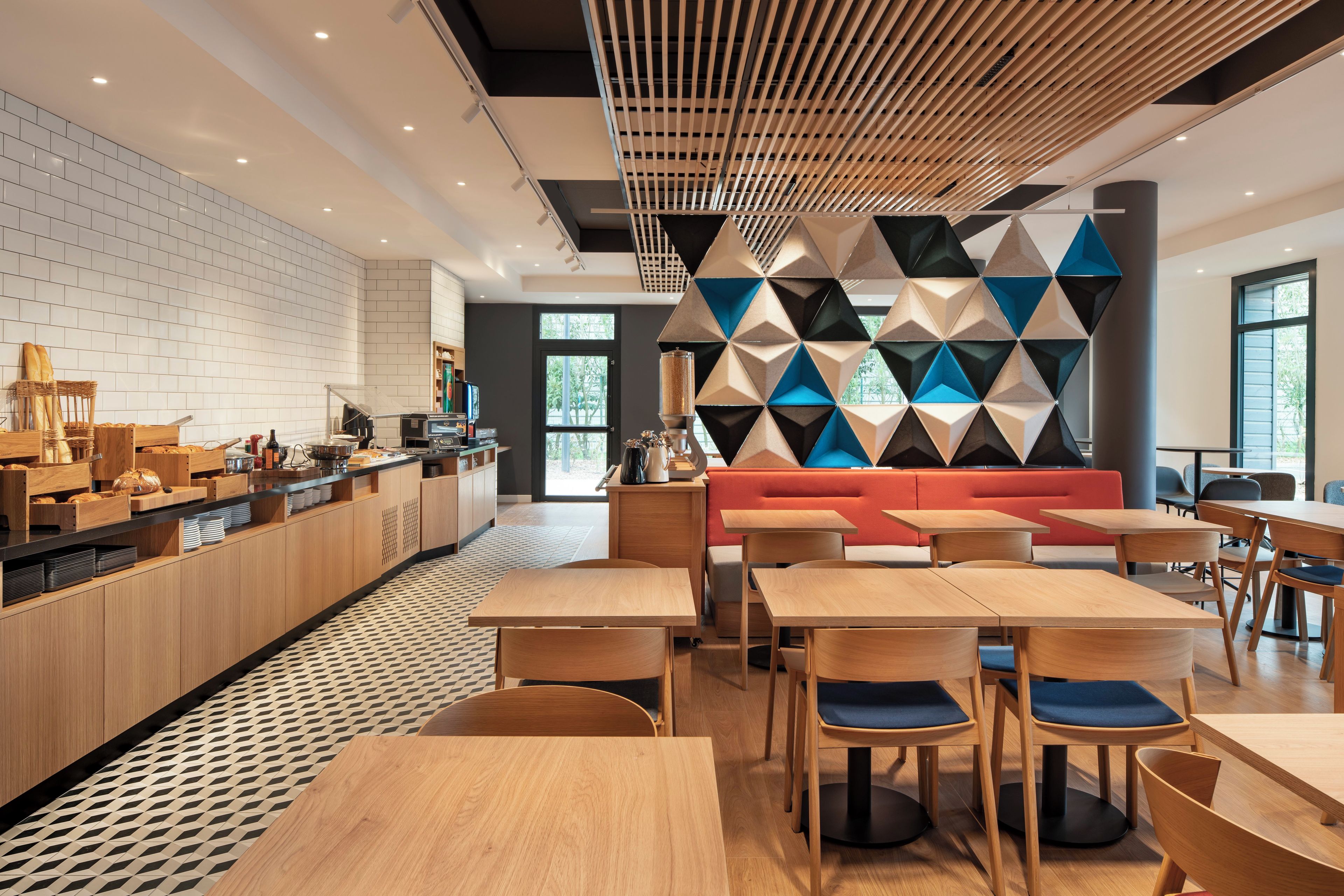 A modern cafe with wooden tables and chairs, a red sofa, and a geometric wall design.