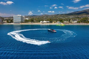 Une scène côtière avec un bateau laissant une vague derrière lui, une plage avec des parasols et un bâtiment en arrière-plan.