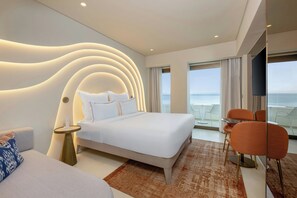 Habitación de hotel moderna con una cama grande, un área de descanso con sillas naranjas, un televisor y vista al mar.