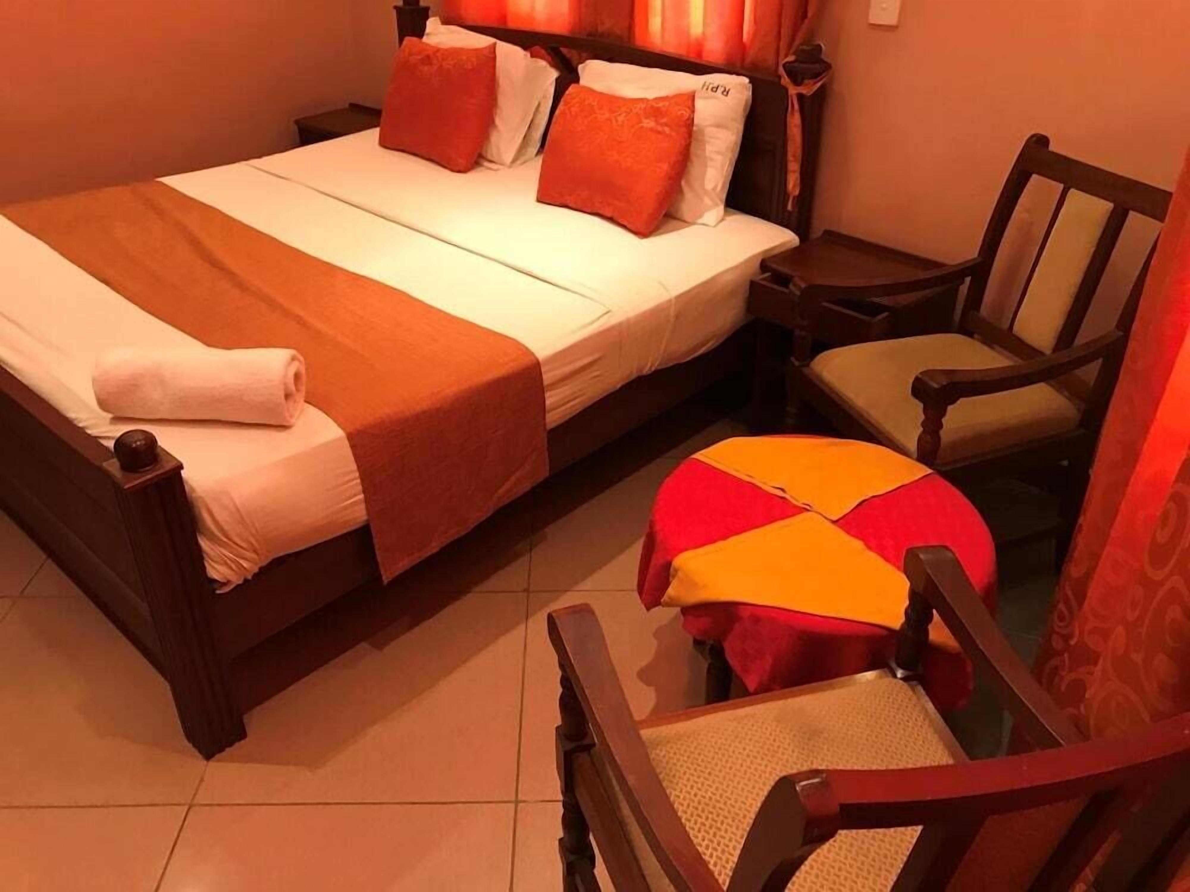 A neatly made bed with a white and orange bedspread, two pillows, and a rolled white towel. There is a wooden chair with a cushion and a small round table with a red and orange cushion.