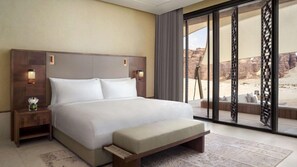 A modern hotel room with a large bed, a nightstand, a bench, and a view of a desert landscape through large windows. - Banyan Tree Alula (Al-'Ula)