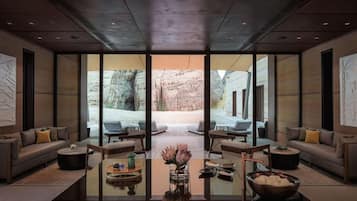 A modern hotel lobby with a large glass wall offering a view of a rocky outdoor area. There are seating areas with sofas and tables, and a central coffee table with decorative items.