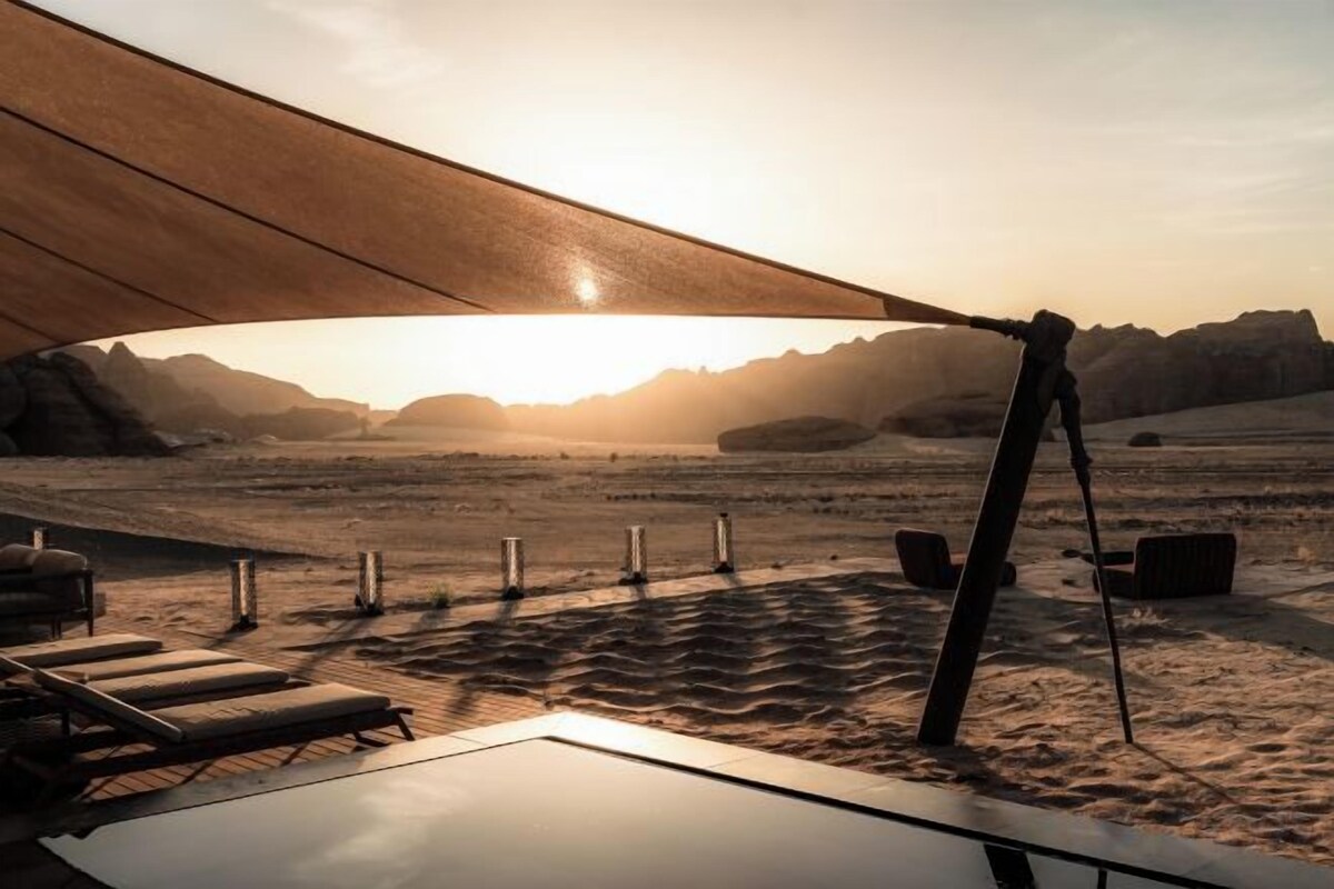 A desert landscape with a sunlit sky, a tent-like structure, and a series of lounge chairs.