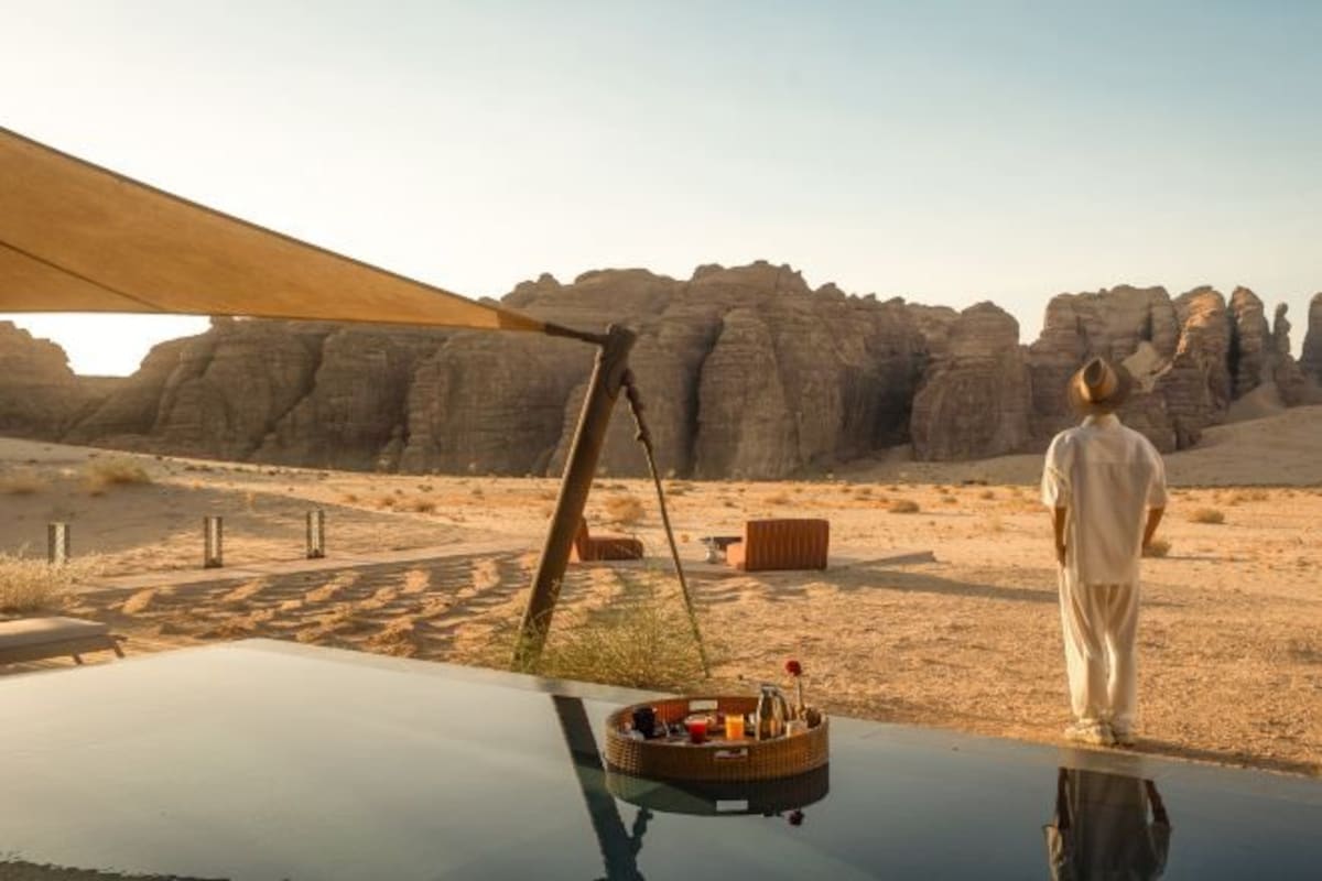 A desert landscape with a person standing by a vehicle, a large rock formation in the background, and a clear sky.