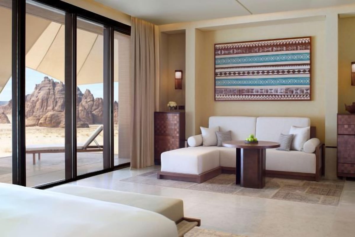 A hotel room with a bed, a sofa, a small table, and a view of a desert landscape through large windows.
