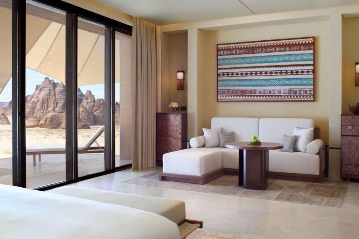 A hotel room with a bed, a sofa, a small table, and a view of a desert landscape through large windows.
