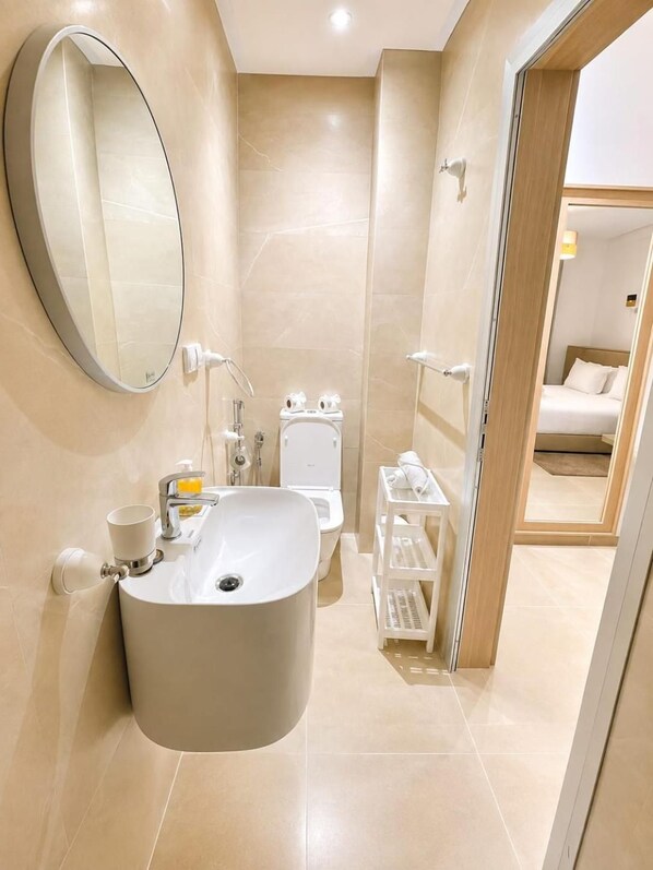 A modern bathroom with a large oval mirror, a white sink, a toilet, and a small shelf with toiletries. - Sables D'or Appart & Hotel (Agadir)