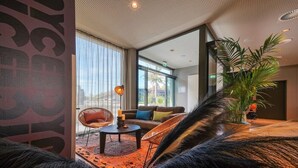 Eine moderne Hotelhalle mit großem Fenster, einer Couch, Sesseln, einem Couchtisch und einer Topfpflanze.