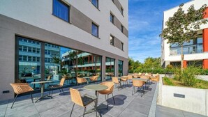 A modern outdoor seating area with wicker chairs and tables, large windows, and a clear sky. - Nyce Hotel Ulm (Ulm)