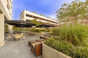 Un espacio de estar al aire libre con mesas y sillas, sombrillas y vista a un edificio al fondo.