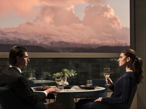Una mesa de comedor con comida, copas de vino y vistas de montañas a través de una ventana.