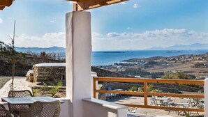 A terrace with stone walls, wicker furniture, and a view of the sea and distant hills. - Dolce Vista - Sea View Apartments (Paros)