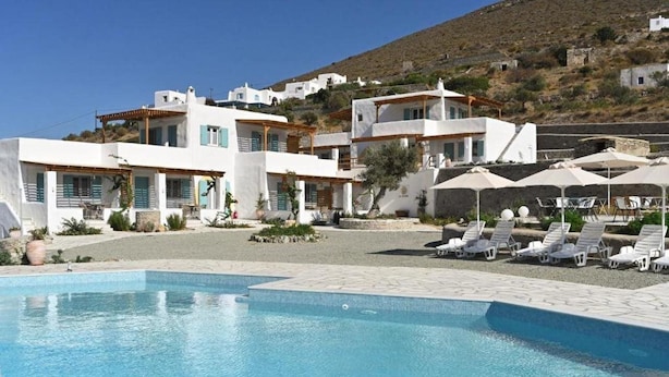 A pool area with lounge chairs and umbrellas, surrounded by white buildings with blue shutters, set against a hillside backdrop. - Dolce Vista - Sea View Apartments (Paros)