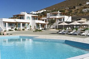 A pool area with lounge chairs and umbrellas, surrounded by white buildings with blue shutters, set against a hillside backdrop. - Dolce Vista - Sea View Apartments (Paros)