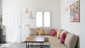 A living room with a sofa, a wooden table, a ceiling fan, a window with curtains, a painting on the wall, and a door. - Dolce Vista - Sea View Apartments (Paros)