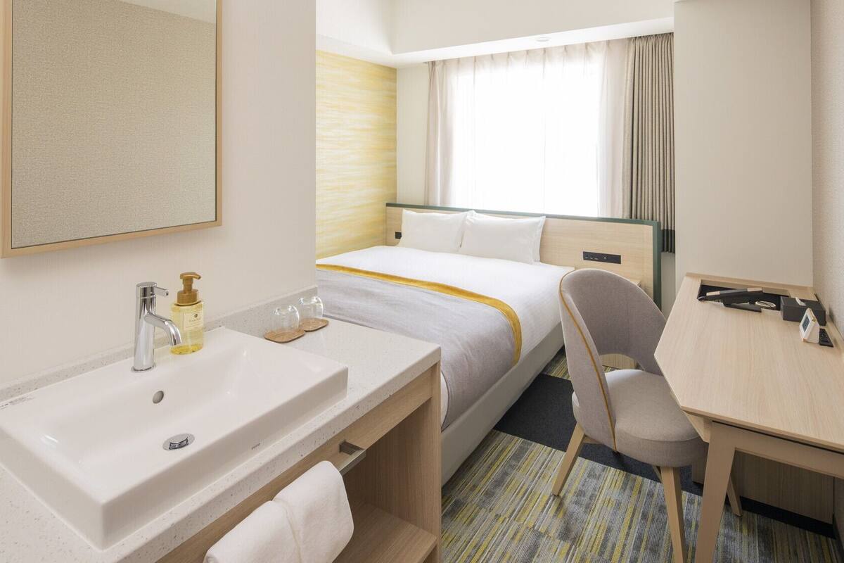 A hotel room with a bed, a desk, a chair, a sink, and a mirror.