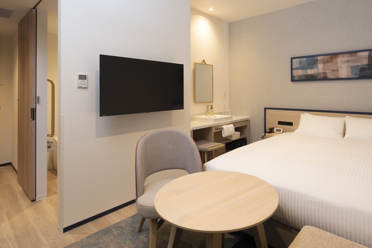 A hotel room with a bed, a desk, a chair, a TV, a bathroom, and a painting on the wall.