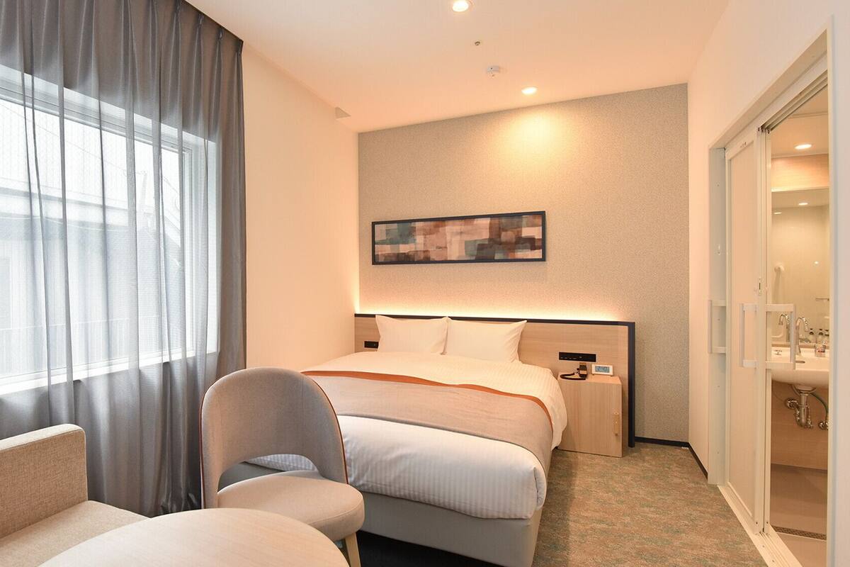 A hotel room with a bed, a chair, a nightstand, a painting on the wall, and a bathroom visible through an open door.