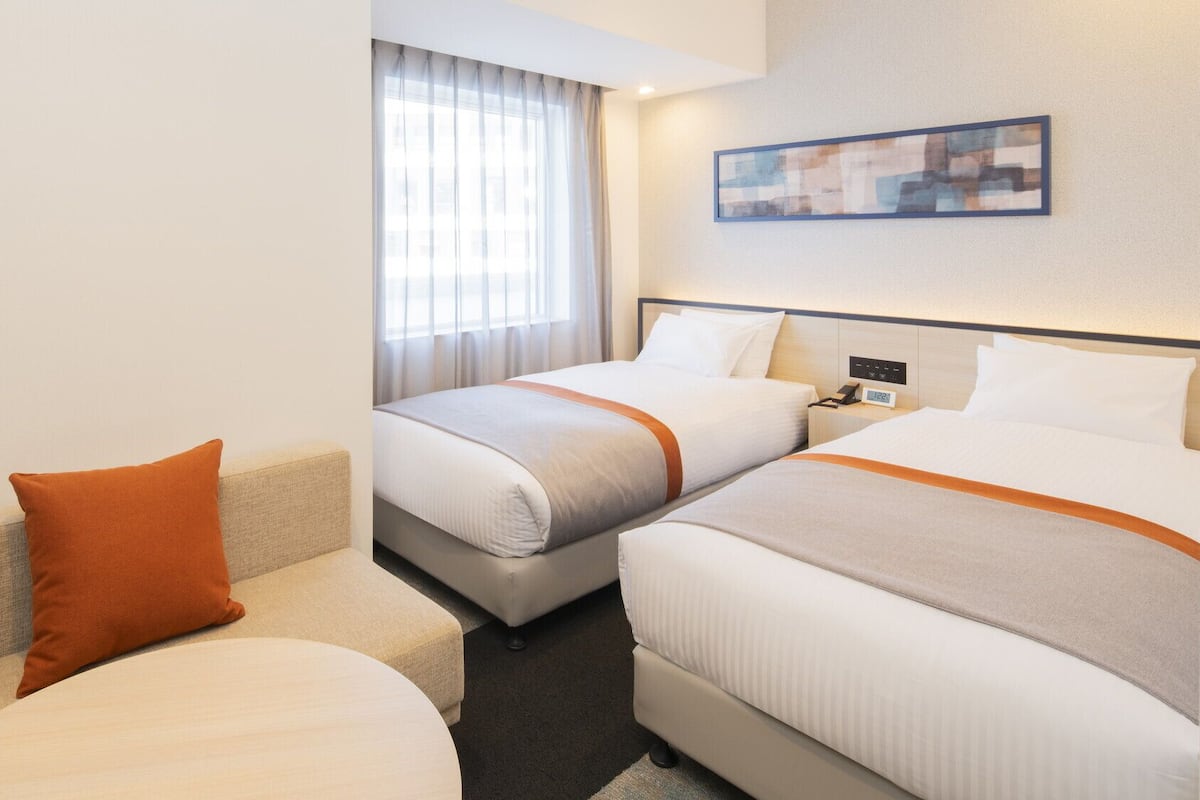 A hotel room with two beds, a sofa, a window with curtains, a painting on the wall, and a bedside table with a phone and clock.