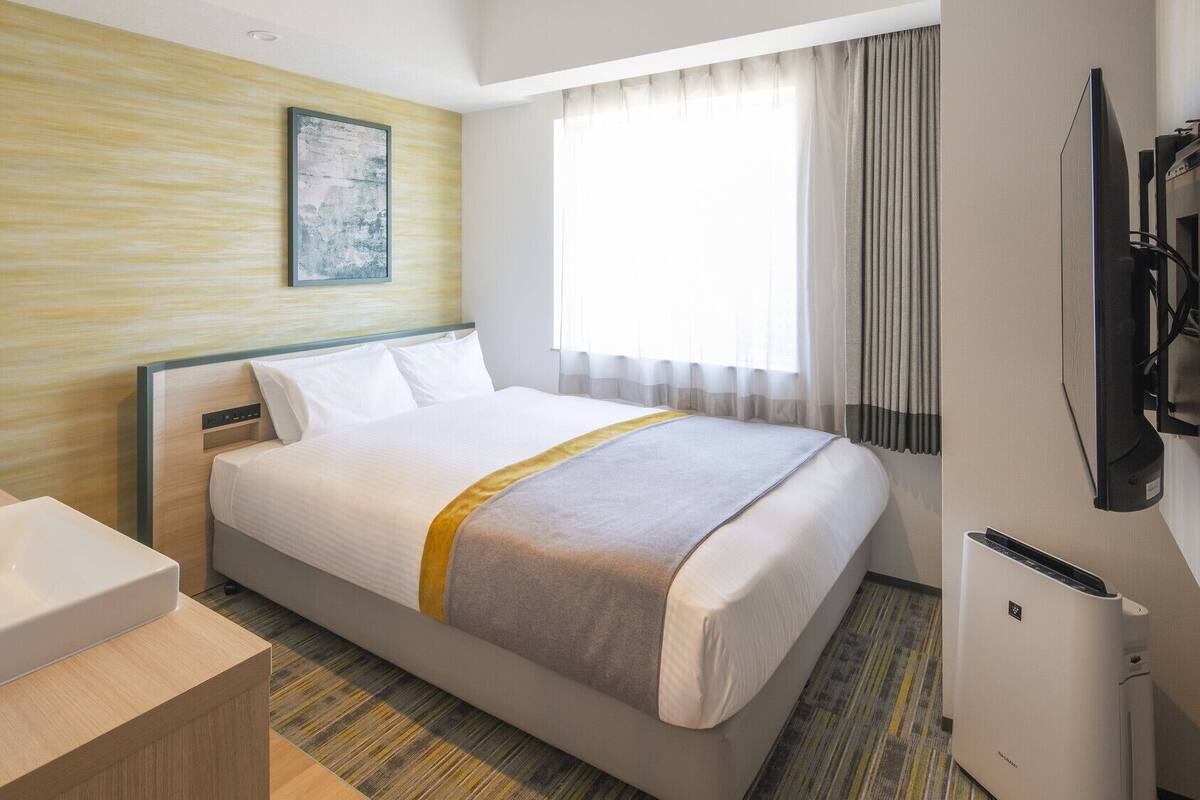 A hotel room with a bed, a bedside table with a sink, a television, and a window with curtains.