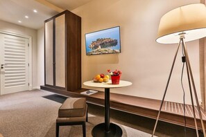 Ett hotellrum med en säng, ett nattduksbord med en lampa, ett litet bord med frukt och blommor, en väggmonterad TV och en dörr.