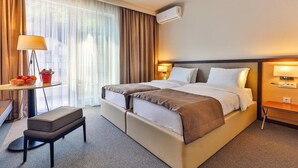 A hotel room with a large bed, bedside lamps, a desk with a lamp, a chair, and a window with curtains. - Bono Hotel (Bečići)