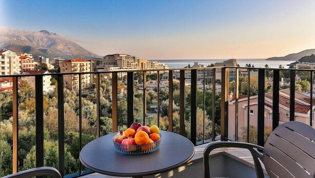 A balcony with a table and chairs, a bowl of fruit, and a view of a coastal cityscape with buildings and mountains in the background. - Bono Hotel (Bečići)