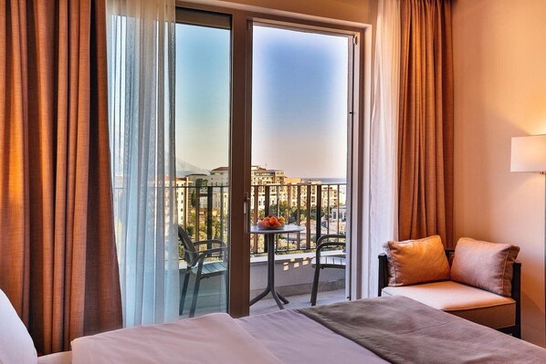 A hotel room with a bed, a small table with fruit, a chair, and a balcony with a view of buildings and mountains. - Bono Hotel (Bečići)