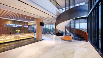 A modern hotel lobby with a marble floor, a curved staircase, and a reception desk with a sign.