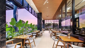 A modern indoor dining area with wooden tables and chairs, large windows, and indoor plants. - Harper Kupang (Kupang)