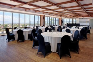 A conference room with round tables, black chairs, and a large window with a city view. - Harper Kupang (Kupang)