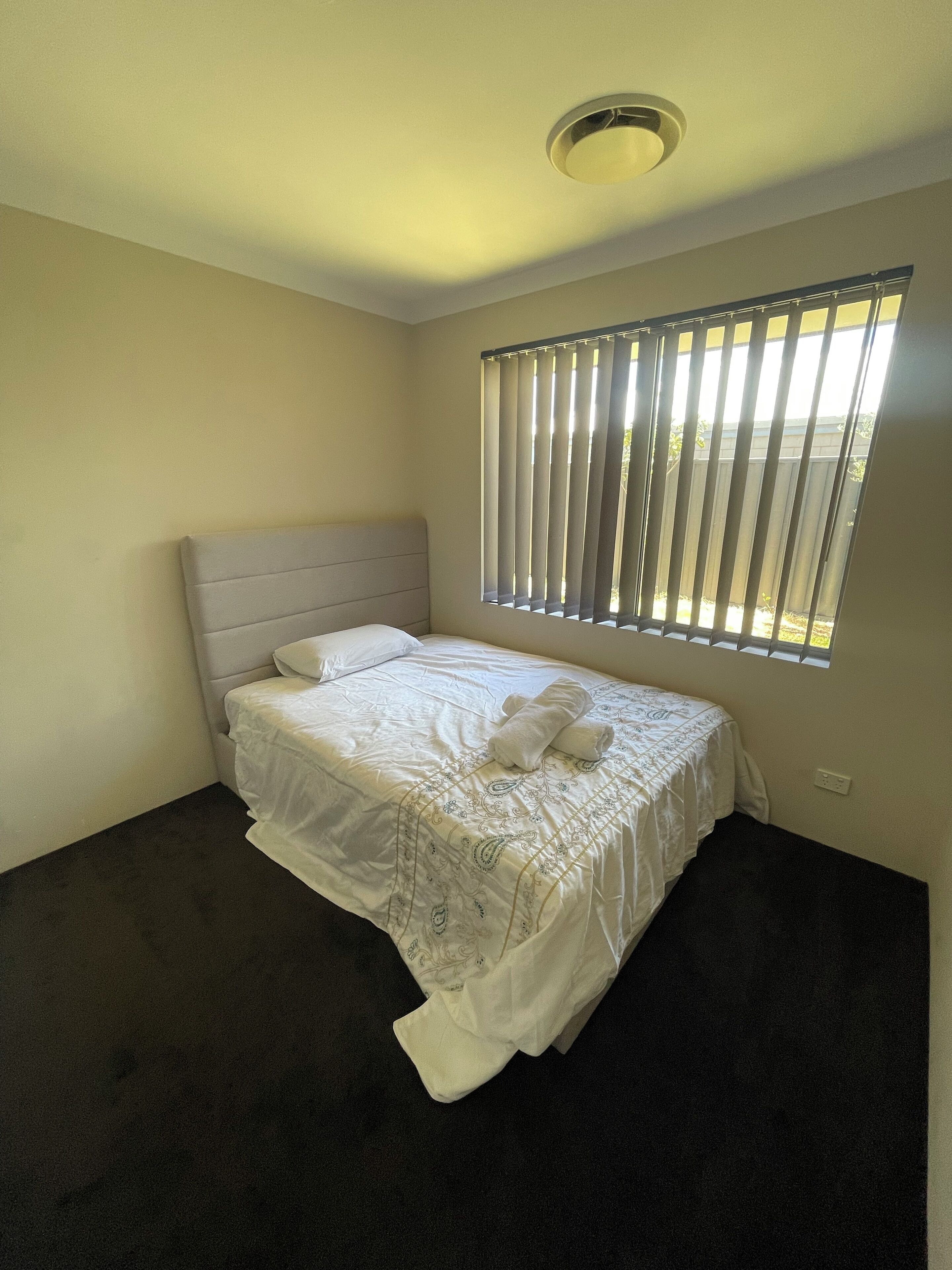 A neatly made bed with a headboard, a window with vertical blinds, and a light switch on the wall.