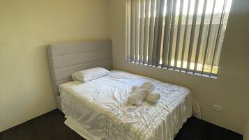 A neatly made bed with a headboard, a window with vertical blinds, and a light switch on the wall.