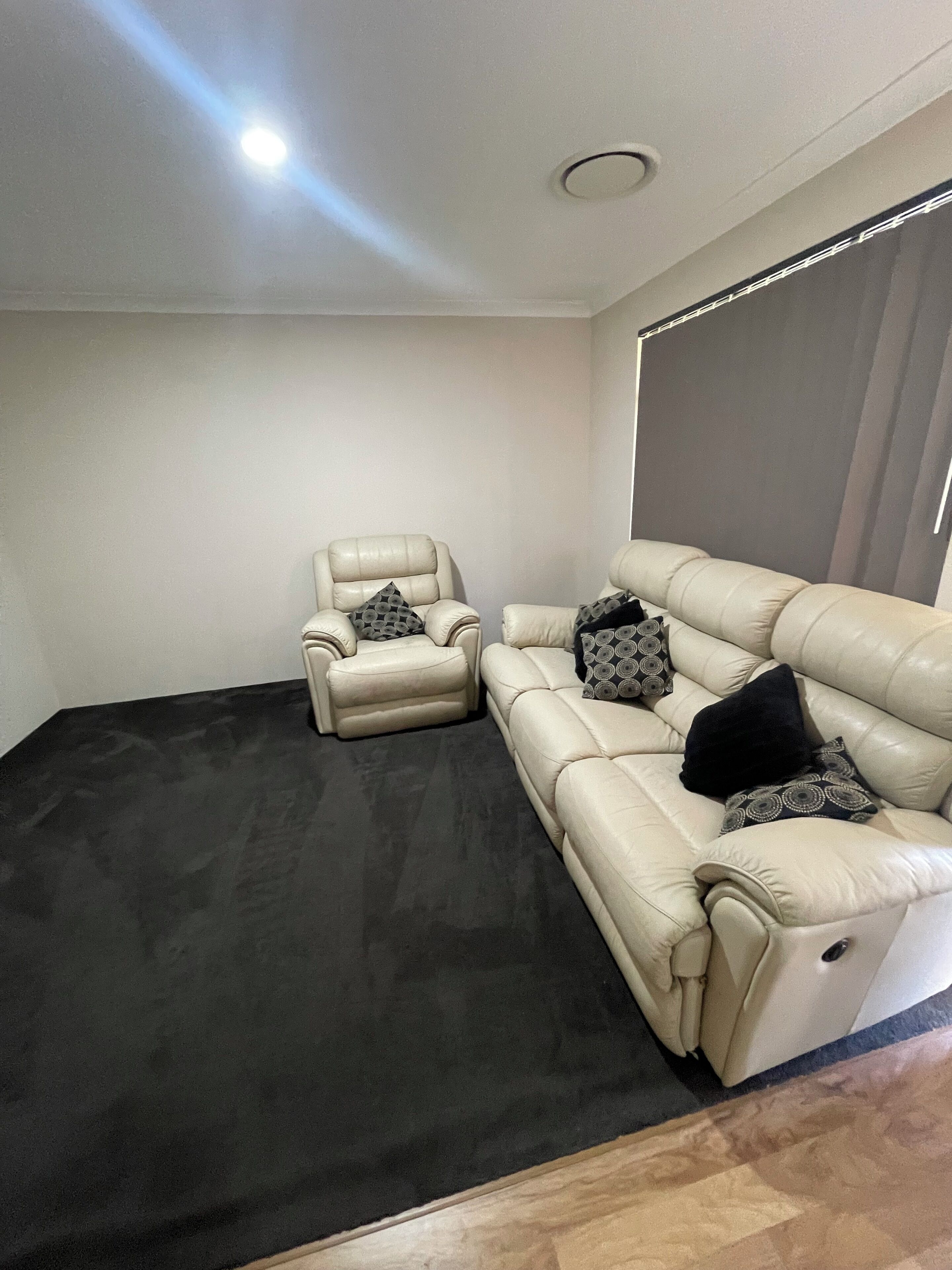 A room with a black carpet, a beige sofa, a leather armchair, and a patterned pillow.