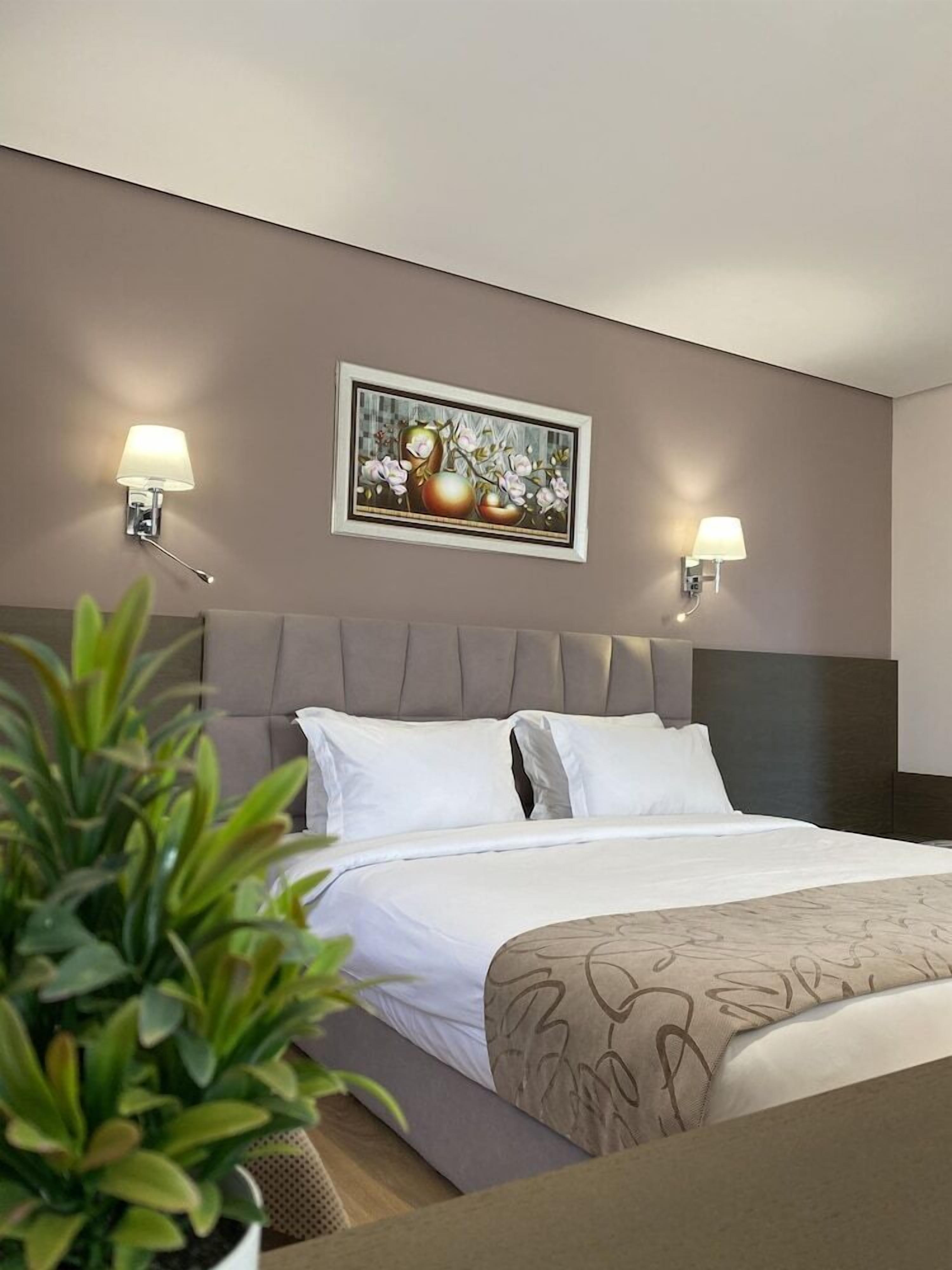 A hotel room with a bed, two wall-mounted lamps, a framed picture above the bed, and a potted plant in the foreground.
