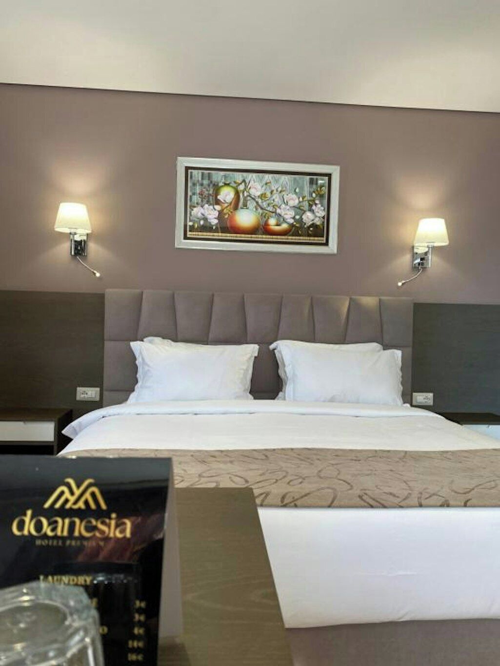 A hotel room with a bed, two bedside lamps, a framed artwork above the bed, and a visible logo on the bedspread.