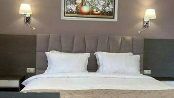 A hotel room with a bed, two bedside lamps, a framed artwork above the bed, and a visible logo on the bedspread.