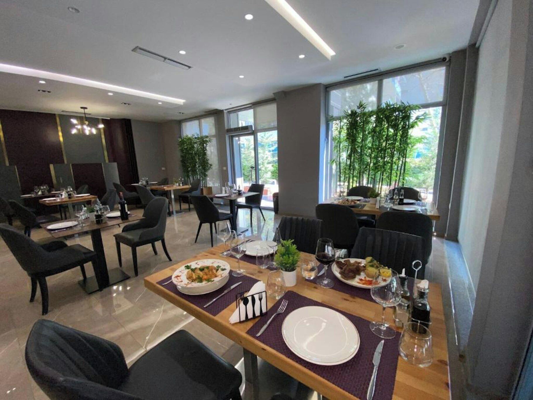 A modern dining area with multiple tables and chairs, large windows with a view of greenery, and a clean, contemporary design.