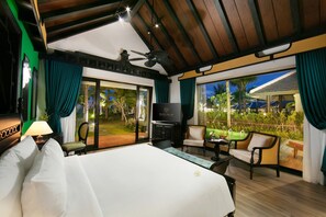 Ein Hotelzimmer mit einem großen Bett, einem Fernseher, einem Schreibtisch mit Stuhl und Blick auf einen Garten und Palmen.