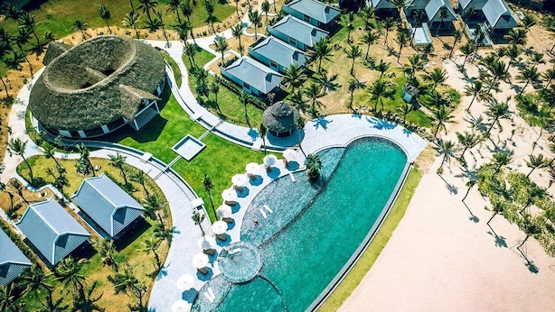 Vista aérea de un complejo turístico con una piscina central, varias cabañas y un edificio circular de techo de paja.